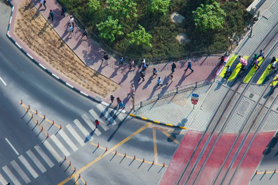 Overhead view of doxens of commuters walking on sidewalk and crossing street in full sun.